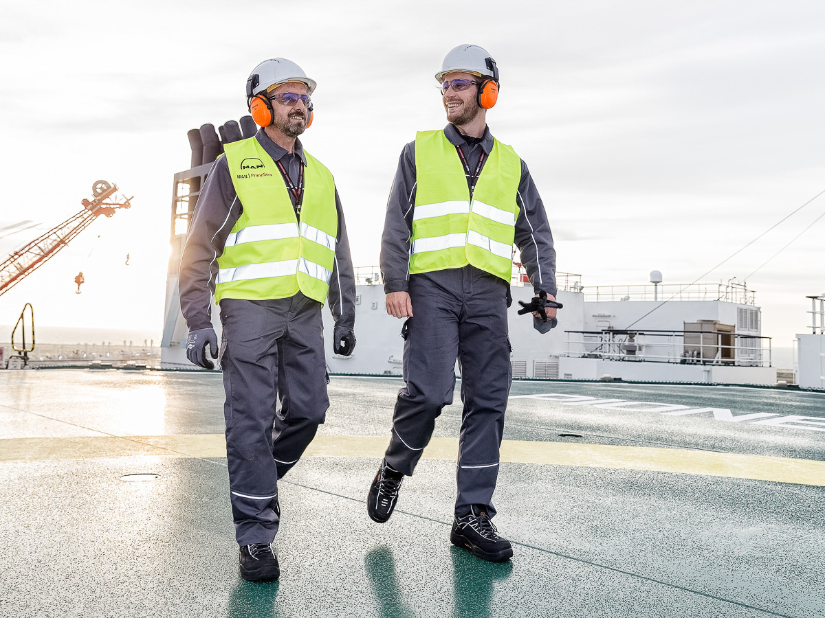 Zwei Männer in Sicherheitskleidung, Helme, Warnwesten und Gehörschutz, die auf einem Schiff arbeiten.