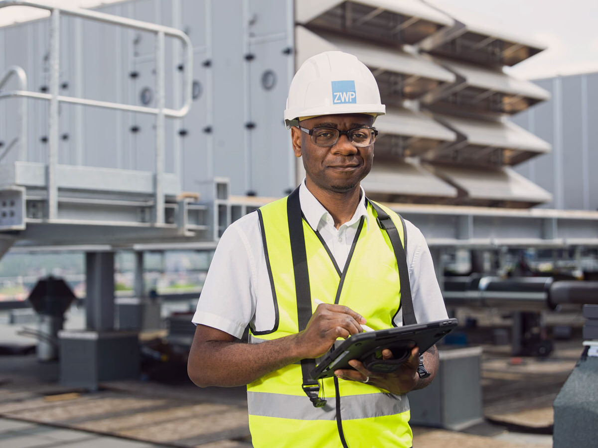 Ingenieur auf Baustelle – Arbeiter mit Helm und Sicherheitsweste nutzt ein Tablet, vor einer technischen Anlage.