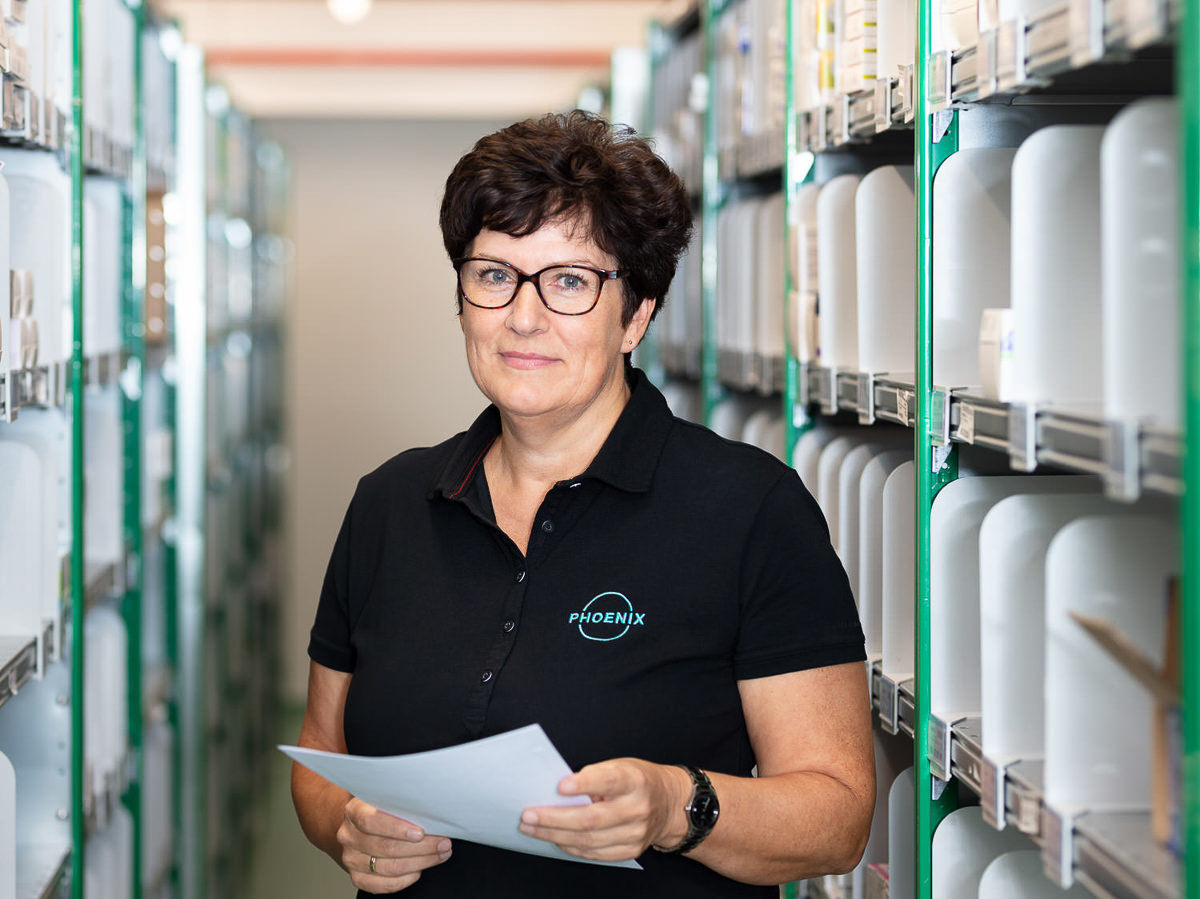 Mitarbeiterin in einem Logistikzentrum in München, umgeben von Regalen, hält ein Dokument und schaut in die Kamera.