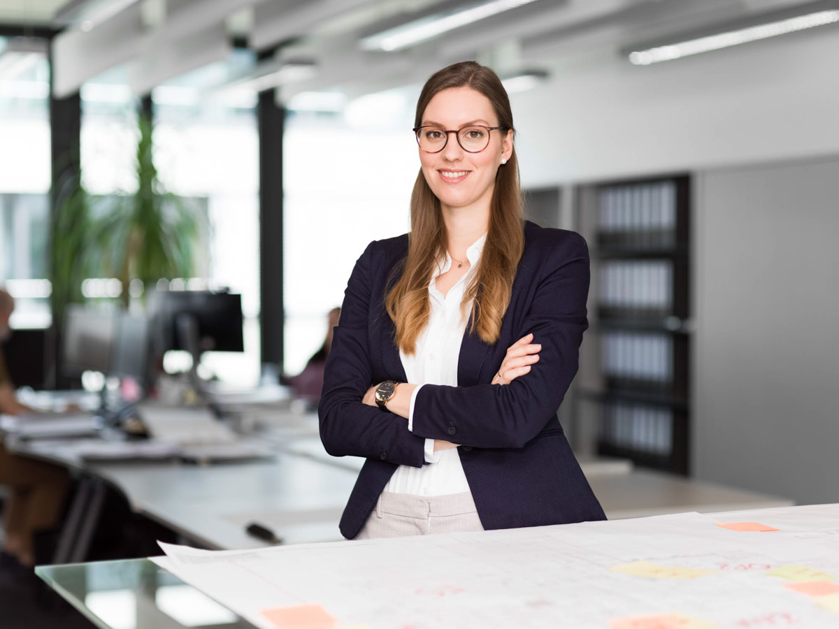 Geschäftsfrau mit verschränkten Armen in modernem Büro, lächelnd vor Arbeitsunterlagen.